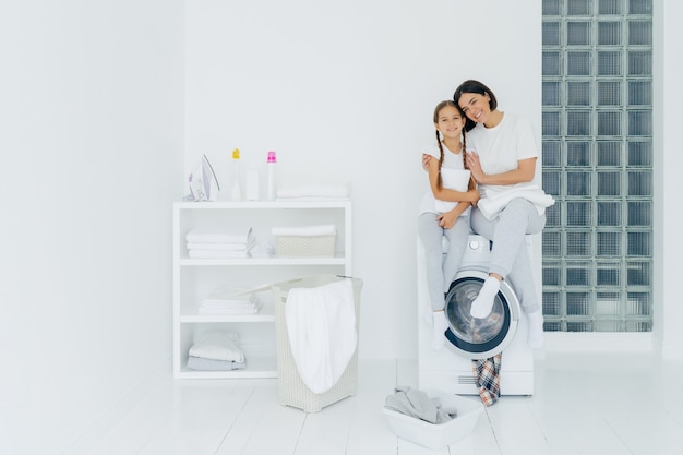 Geschoten van mooie vrouw en zijn kleine dochter omhelzen en glimlachen aangenaam, zitten op wasmachine, wassen linnen in wasruimte, hebben vriendschappelijke relatie, doen was thuis. huishoudelijk werk concept