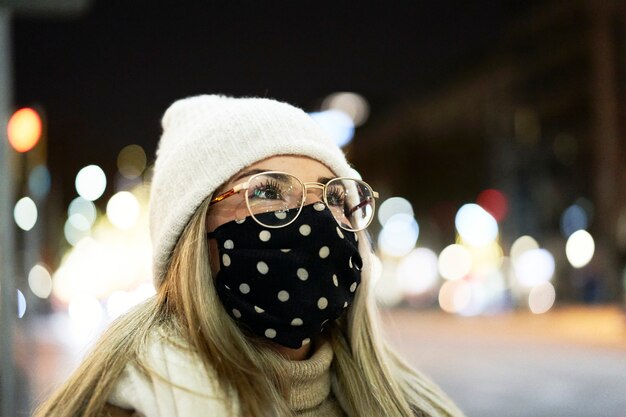 Gesloten shot van een jonge blonde vrouw met een masker die 's nachts rondkijkt in een stad, met veel lichten op de achtergrond. winter omgeving.