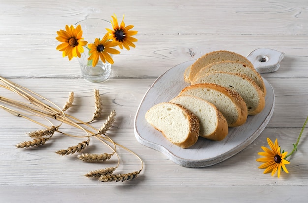 Gesneden brood op snijplank, aartjes van tarwe en gele bloemen in glas op witte houten tafel. Rustieke stijl.