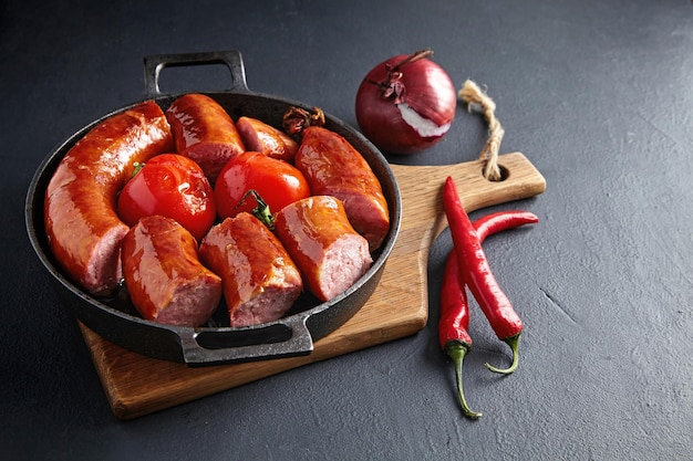 Gesneden gebakken varkensworst in een gietijzeren pan met tomaten op een houten snijplank op een zwarte stenen tafel