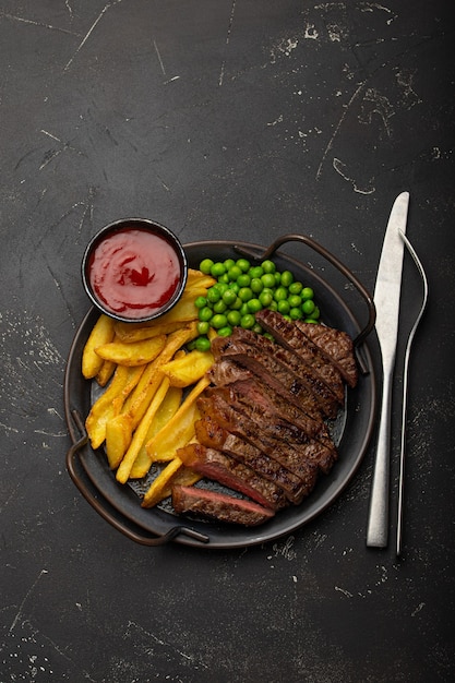 Gesneden gegrild vlees biefstuk met chips, frietjes en groene erwten geserveerd met rode tomatensaus op rustieke metalen plaat op zwarte stenen achtergrond tafel van bovenaf