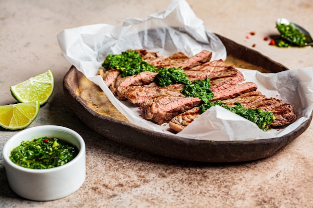 Gesneden gegrilde biefstuk met chimichurri saus op een donkere schotel, donkere achtergrond.