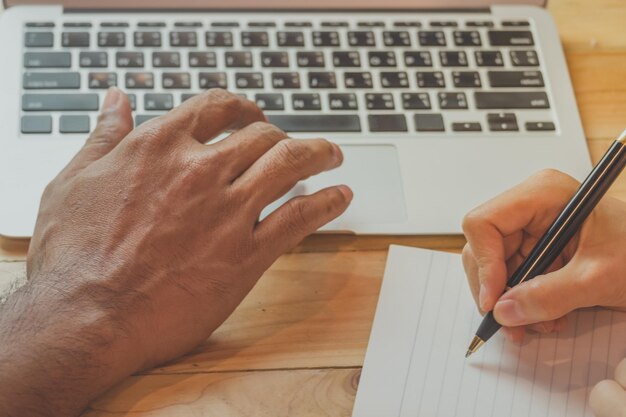 Foto gesneden hand van een man die een laptop gebruikt door een vrouw die in een boek op tafel schrijft