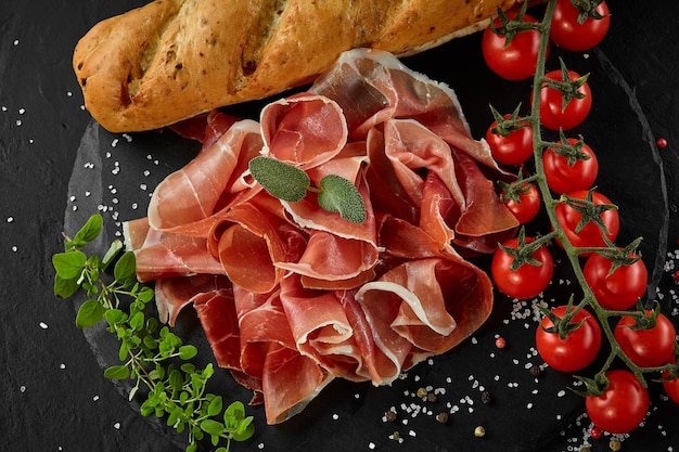 Gesneden jamon kerstomaatjes stokbrood kruiden en specerijen op zwarte steen leisteen bord tegen een donkergrijze achtergrond Close-up shot Bovenaanzicht