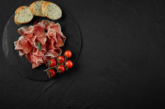 Gesneden jamon kerstomaatjes stokbrood kruiden op zwarte steen leisteen bord tegen een donkergrijze achtergrond Close-up shot Bovenaanzicht Kopieer ruimte