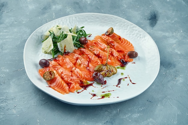 Gesneden licht gezouten zalm met rucola en parmezaan. Rode vissashimi met kappertjes en olijven op een witte plaat. Vers zee-eten