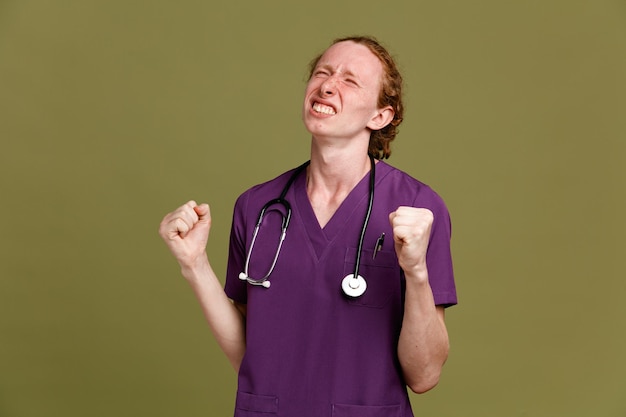 Gespannen tonen ja gebaar jonge mannelijke arts dragen uniform met stethoscoop geïsoleerd op groene achtergrond