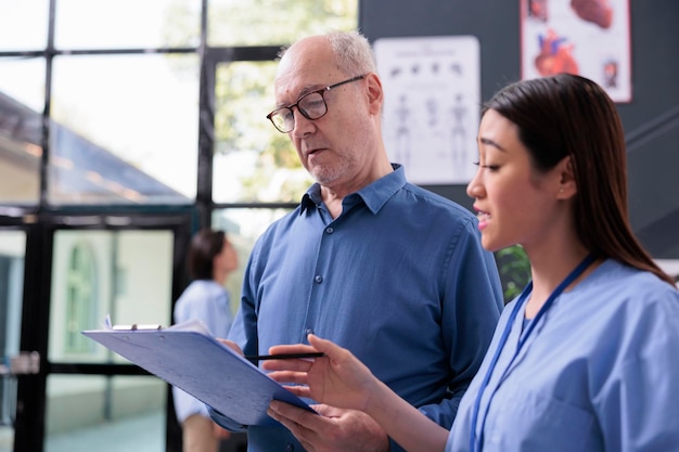 Gespecialiseerde assistent die aan de oude patiënt laat zien waar het medische verzekeringsdocument moet worden ondertekend voordat het onderzoek in de ziekenhuisreceptie begint. Verpleegkundige die ziektesymptomen uitlegt en behandeling bespreekt