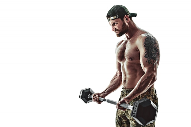 Gespierde atleet bodybuilder man in camouflage broek met een naakte torso training met halter op een witte muur.
