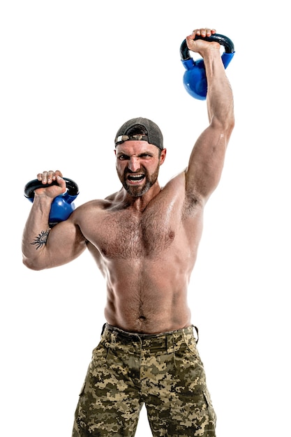 Gespierde atleet bodybuilder man in camouflage broek met een naakte torso training met kettlebell op een witte muur. Isoleren