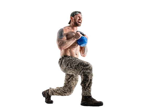 Gespierde atleet bodybuilder man in camouflage broek met een naakte torso training met kettlebell op een witte muur. Isoleren