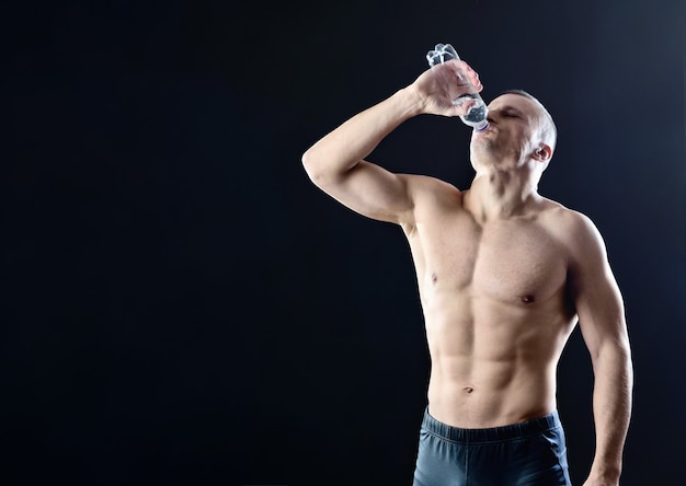 Gespierde atletische man drinkt gretig water uit een doorzichtige plastic fles op een donkere studioachtergrond met achtergrondverlichting.