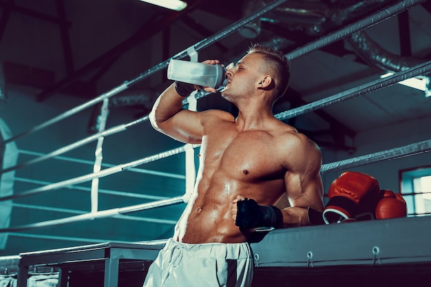 Gespierde blanke man op dieet rusten na boksoefening en drinkwater uit de shaker in de sportschool.