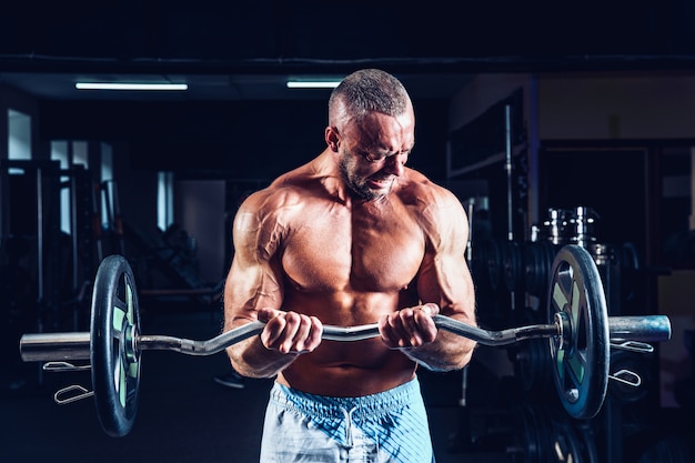 Gespierde bodybuilder man doet oefeningen op biceps met grote halter in sportschool