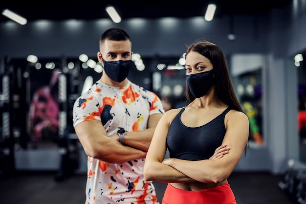 Gespierde fit paar met gezichtsmaskers permanent in sportschool met gekruiste armen. Gezonde gewoonten, coronaviruspreventie, bodybuilding