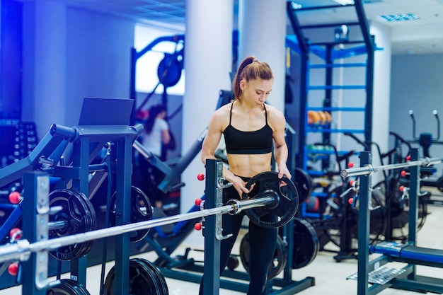 Gespierde fitness vrouw kijkt naar de schijf en zet deze in de barbell in de sportschool Ernstige sterke jonge sportvrouw met schijf bij barbell