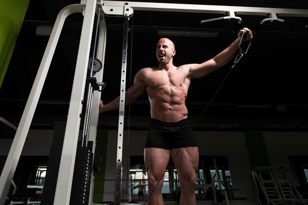 Gespierde fitnessbodybuilder doet zware oefeningen voor schouders op kabelmachine in de sportschool