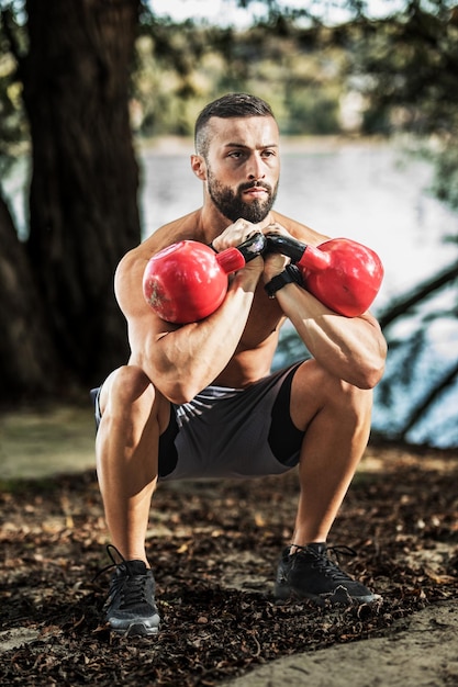 Gespierde jonge man met naakte torso doet squat oefeningen met kettlebell in de natuur, in de buurt van de rivier.