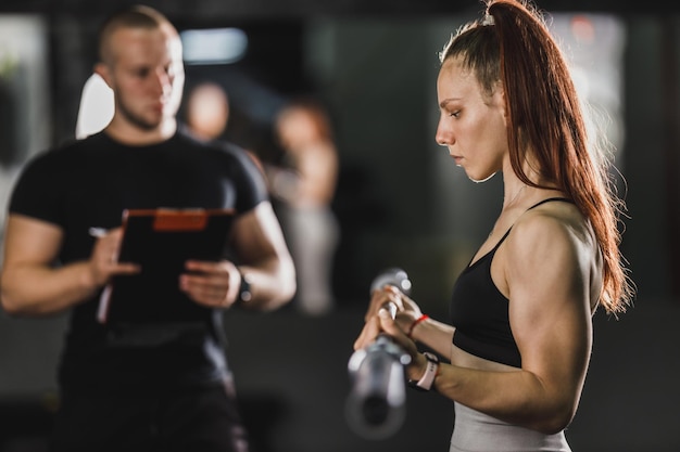 Gespierde jonge vrouw doet krachttraining met coach in de sportschool. Ze pompt haar biceps-spier op met barbell.