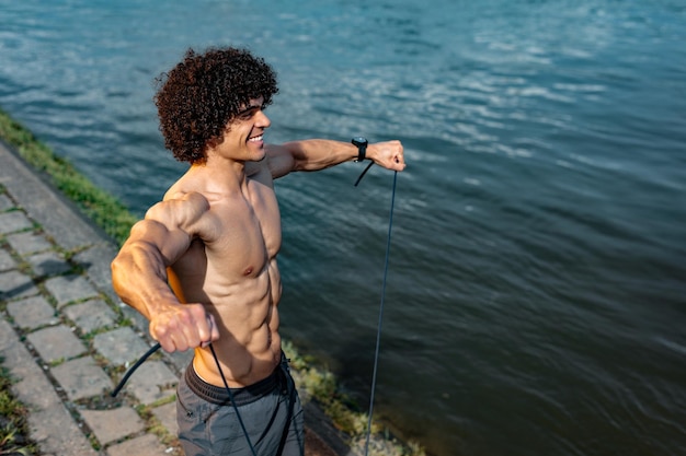 Gespierde jongeman met naakte torso, doet sterke lichaamstraining met elastische rubberen band, bij de rivier.