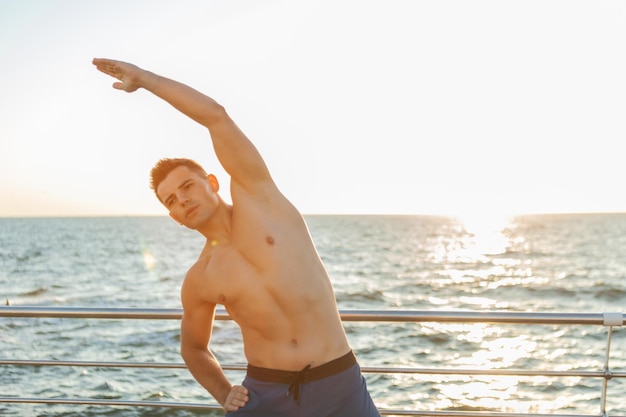 Gespierde knappe man met naakte torso doet warming-up oefening voor training op het strand bij zonsopgang