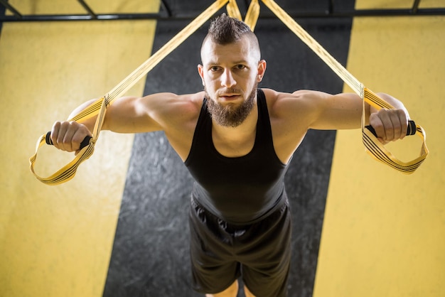 Foto gespierde man doet oefeningen met fitnessriemen in de sportschool met zwart en geel interieur