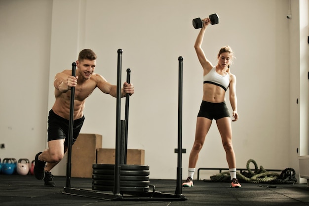 Gespierde man doet slee push-oefening terwijl vrouw handgewichten op de achtergrond opsomt op cross-training in een sportschool Focus ligt op de man