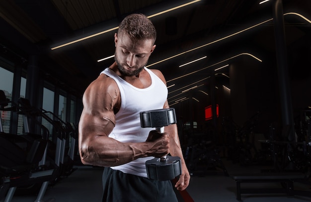 Gespierde man in een wit t-shirt traint in de sportschool met halters. Biceps pompen. Fitness en bodybuilding concept.