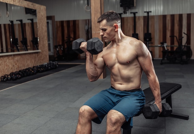 Gespierde man oefent met zware halters, traint biceps terwijl hij op een bankje in de sportschool zit. Bodybuilding en fitness