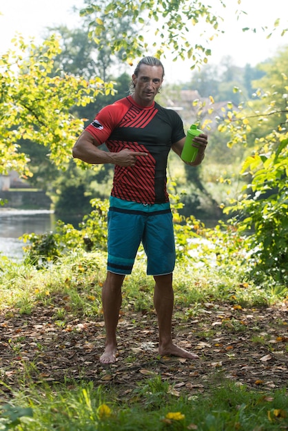 Gespierde man rust na het sporten en drinken uit shaker buitenshuis in de natuur