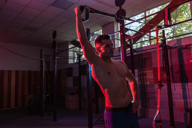 Gespierde man traint met het trx-systeem in een donkere sportschool Atleet met naakte torso-training met weerstandsbanden in roodblauw neonlicht Functionele crosstraining