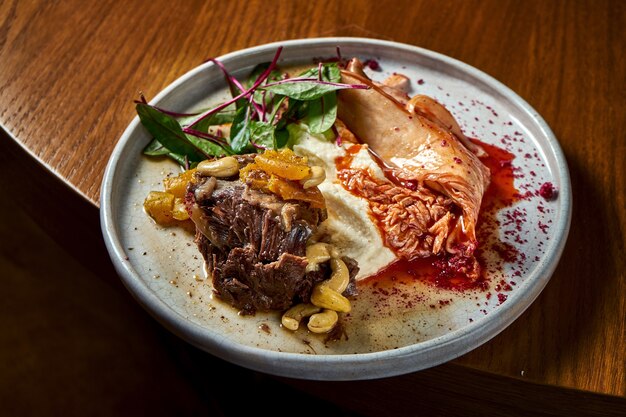 Gestoofde biefstuk met gedroogd fruit en noten gegarneerd met selderij puree en kimchi-kool, geserveerd in een witte plaat op een houten tafel. Restaurant eten