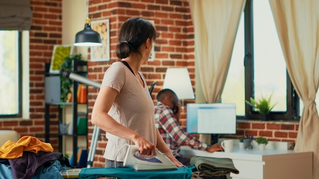 Gestresst persoon gooit blouse naar ontspannen partner, gefrustreerd terwijl ze thuis kleding strijkt. Ongelukkige vrouw die depressief is en probeert appartement schoon te maken, voorjaarsschoonmaak doet.