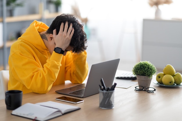 Gestresste Aziatische man zit aan een bureau met een pc die zijn hoofd vastpakt