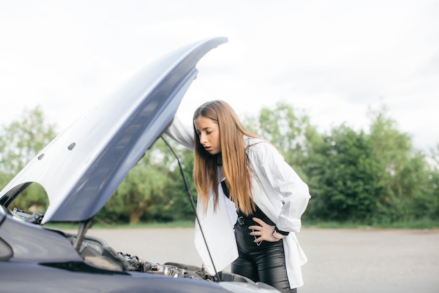 Gestresste vrouw die naar een kapotte auto kijkt en de motor op de landweg in zijaanzicht controleert