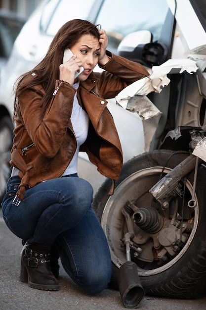 Gestresste vrouw die op mobiele telefoon praat terwijl ze naar haar vernielde auto kijkt na het ongeval