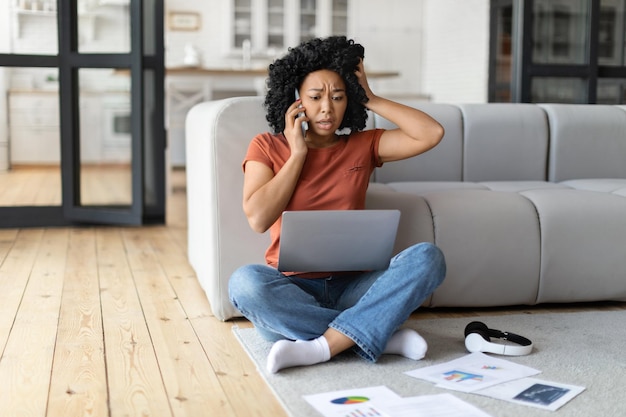 Gestresste zwarte vrouw die op mobiel praat en thuis op laptop werkt