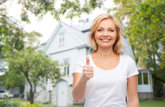 gesture, advertisement, real estate, home and people concept - smiling woman in blank white t-shirt showing thumbs up over private house background