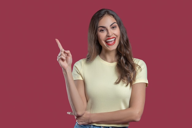 Gesture, attention. Happy young beautiful woman with long wavy hair and bright red lipstick on lips raising hand up and pointing finger