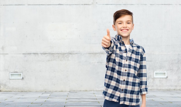 Photo gesture, childhood, gender, fashion and people concept - smiling boy in checkered shirt and jeans showing thumbs up over urban street background