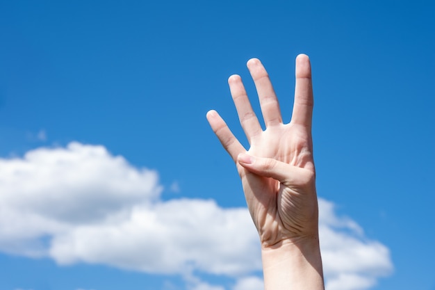 Gesture closeup of a woman's hand showing four fingers, isolated on a background of blue sky with clouds, sign language symbol number four.