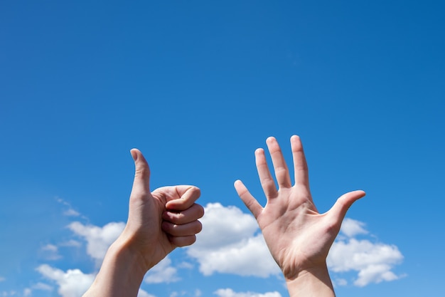 Gesture closeup of a woman's hand showing one opened palm and thumb up isolated on a blue sky background with clouds, sign language symbol number six