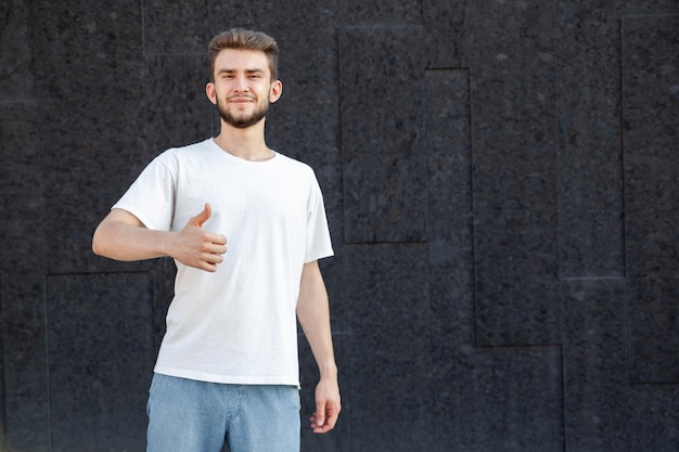 gesture emotion expression and people concept caucasian bearded happy darkhaired man in white tshirt and jeans showing thumbs up with his right hand outside on a dark background with copy space