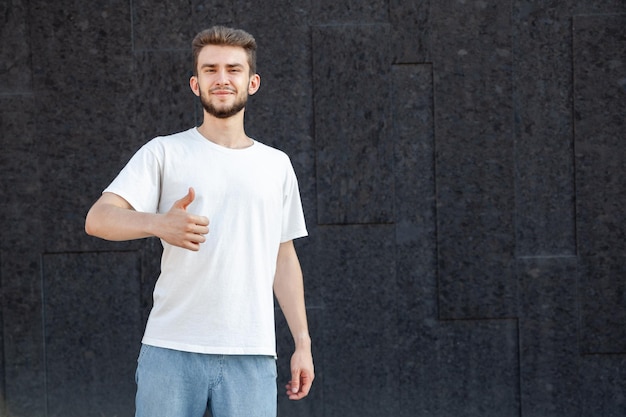 Gesture emotion expression and people concept Caucasian bearded happy darkhaired man in white Tshirt and jeans showing thumbs up with his right hand outside on a dark background with copy space