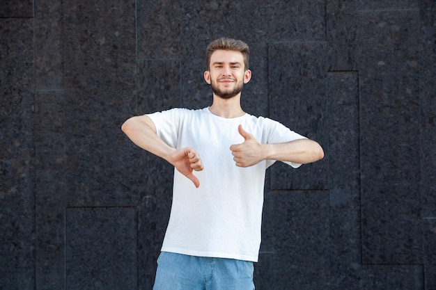 Gesture emotion expression and people concept Caucasian bearded smiling man in white tshirt and jeans showing the likes and dislikes outdoors on a dark background with copy space