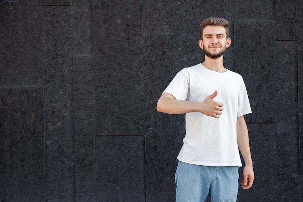 gesture emotion expression and people concept european bearded happy darkhaired man in white tshirt and jeans showing thumbs up with right hand in the street on a dark background with copy space
