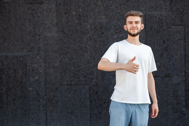 Gesture emotion expression and people concept European bearded happy darkhaired man in white Tshirt and jeans showing thumbs up with right hand in the street on a dark background with copy space