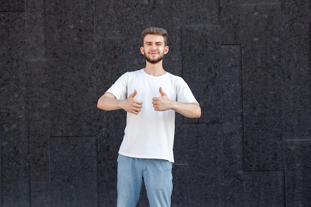 Gesture expression and people concept Caucasian bearded happy darkhaired man in white Tshirt and jeans showing thumbs up with right hand in the street on a dark background with copy space