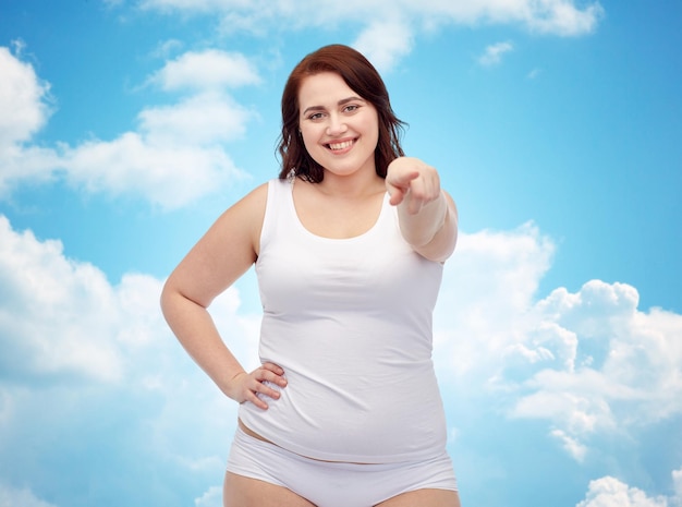 жест, потеря веса и концепция людей - улыбающаяся молодая женщина больших размеров в нижнем белье, показывающая на фоне голубого неба и облаков