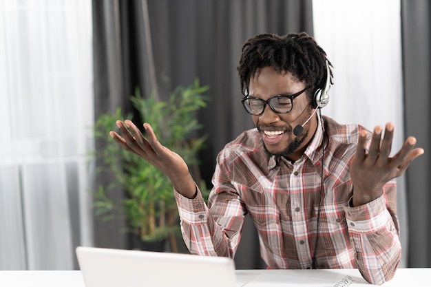 Gesturing afroamericano freelance guardando il laptop con le cuffie durante una videochiamata di gruppo che lavora a distanza a casa